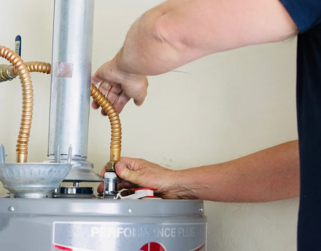 A plumber's hands connect a flexible copper pipe to the top of a silver Performance Plus water heater.