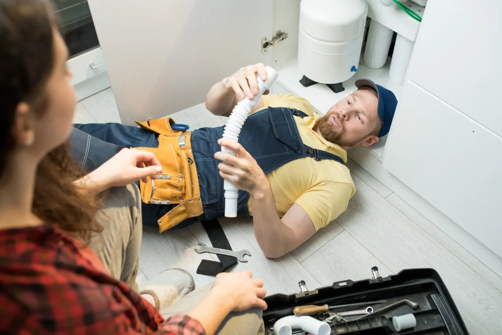 A plumber lying on a kitchen floor shows a pipe to a homeowner sitting nearby.