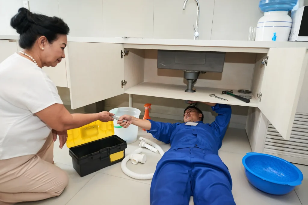 A plumber lying on his back under a sink receives a wrench from a homeowner.