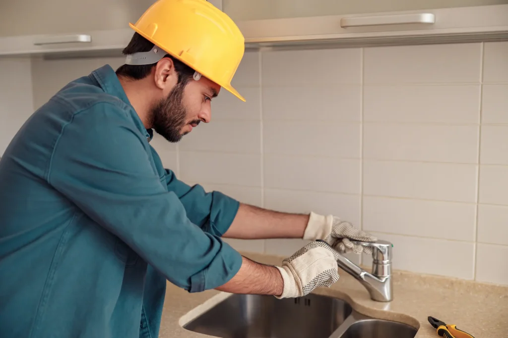 A plumber wearing a hard hat and gloves works on a kitchen sink faucet.