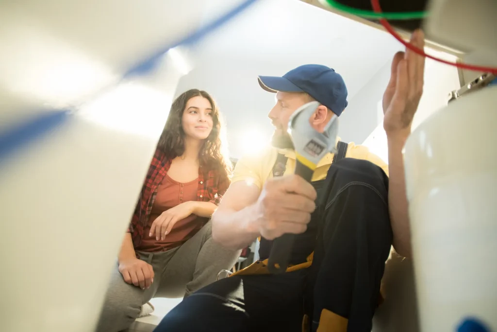 A plumber in a blue cap holds a wrench while talking to a homeowner.