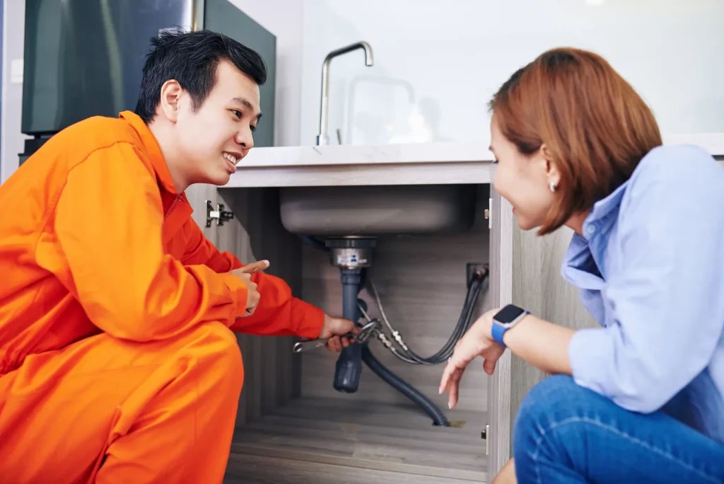 A plumber in orange work clothes explains under-sink plumbing to a homeowner.