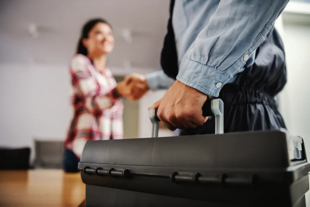 A plumber, carrying a black toolbox, shakes hands with a smiling woman after a job well done.
