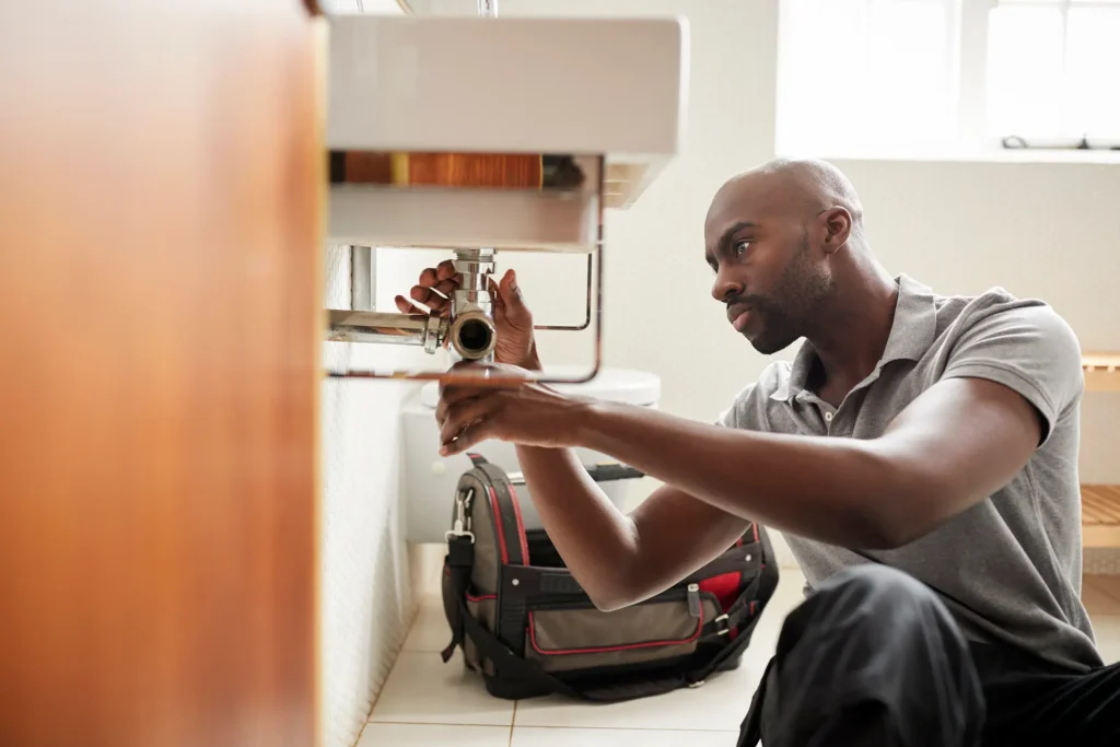 A plumber crouches on a tiled floor, working on pipes under a white sink with a tool bag nearby.