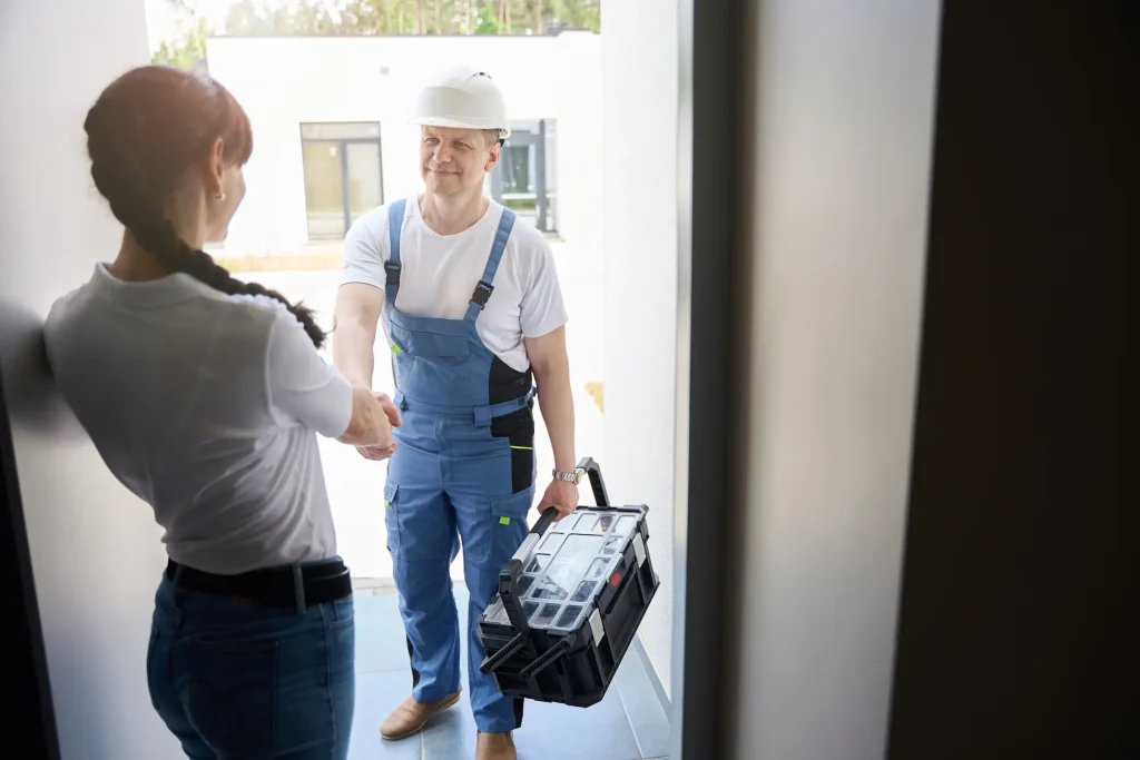 A plumber in a white hard hat and blue overalls shakes hands with a homeowner at a doorway.